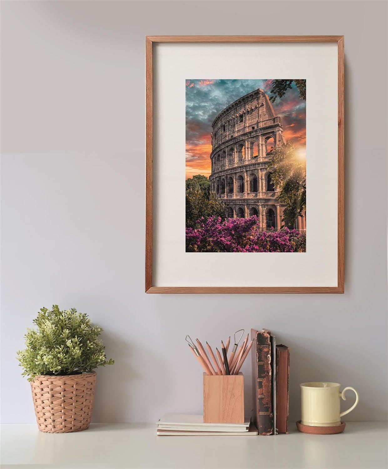 Puzzle Anfiteatro 500 piezas Clementoni montado y enmarcado en madera sobre una pared, con la escena del Coliseo de Roma al atardecer; debajo, una planta, un bote de lápices, libros y una taza.