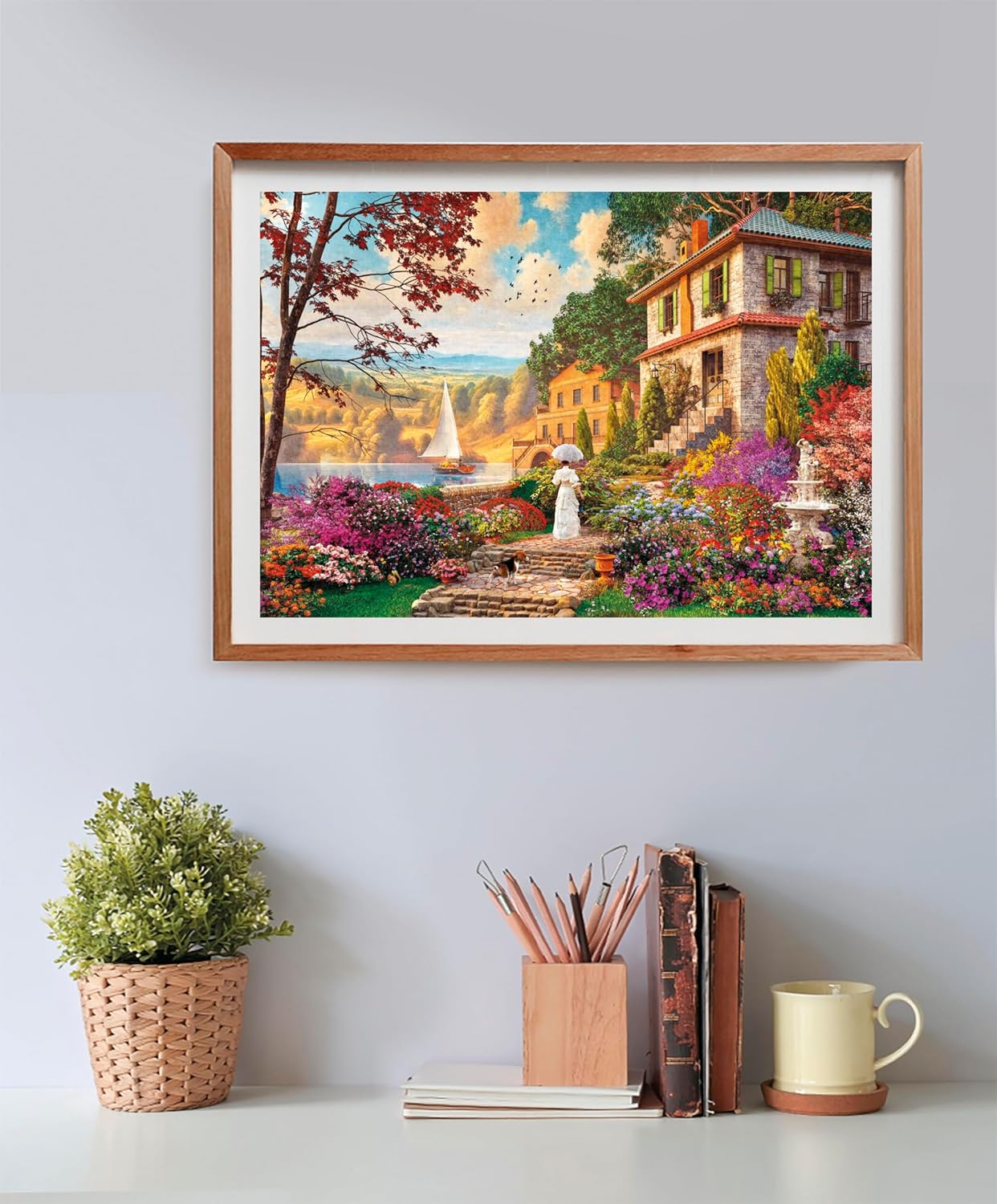 Puzzle Paseo Victoriano 500 piezas Clementoni montado y enmarcado en madera sobre una pared, con la escena de la mujer paseando por el jardín florido junto al lago; debajo, una planta, un bote de lápices, libros y una taza.