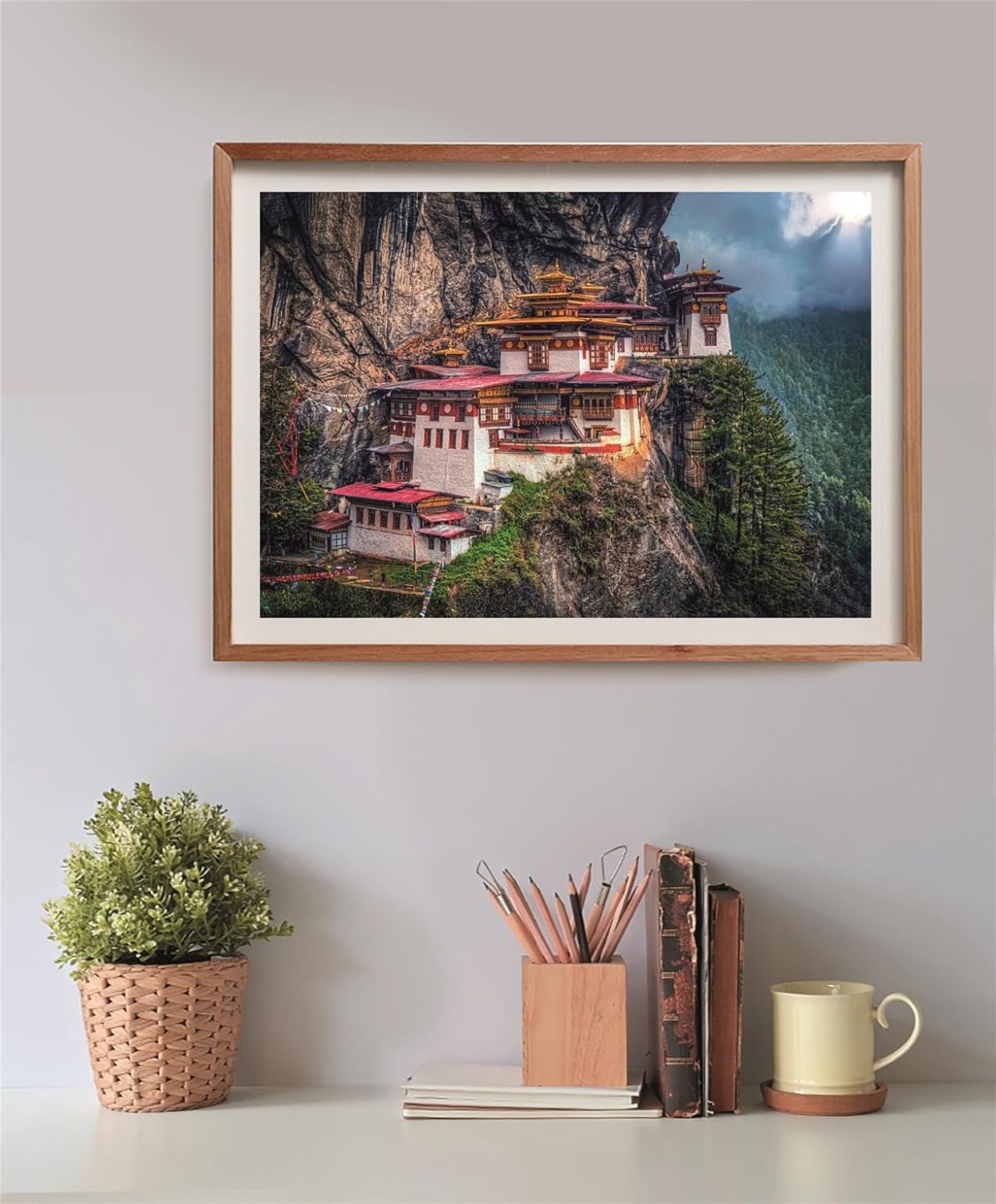 Puzzle El Nido del Tigre 500 piezas Clementoni montado y enmarcado en madera sobre una pared, con la escena del monasterio de Paro Taktsang en el acantilado; debajo, una planta, un bote de lápices, libros y una taza.