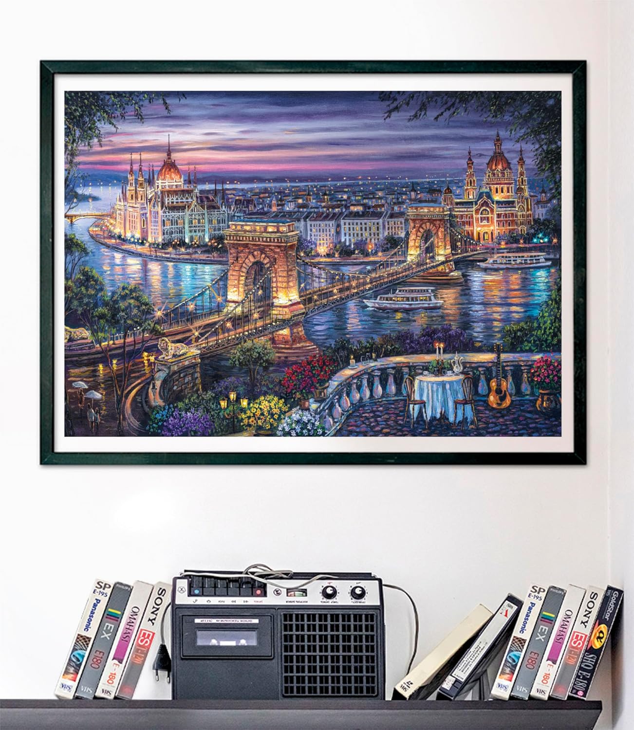 Puzzle Paisaje Nocturno de Budapest 1000 piezas Clementoni montado y enmarcado en negro sobre una pared, con la escena del Puente de las Cadenas al anochecer; debajo, un radiocasete y cintas VHS en una estantería.