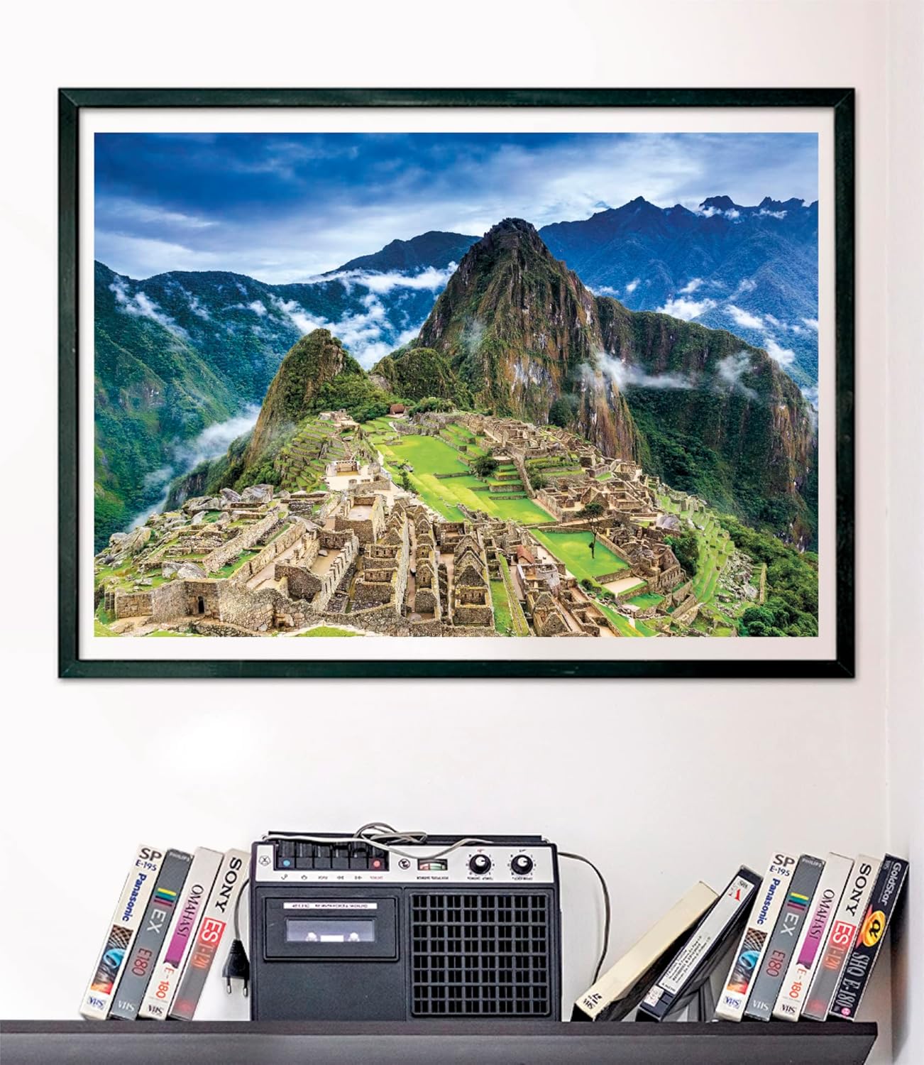 Puzzle Machu Picchu 1000 piezas Clementoni montado y enmarcado en negro sobre una pared, con la vista aérea de la ciudadela inca; debajo, un radiocasete y cintas VHS en una estantería.