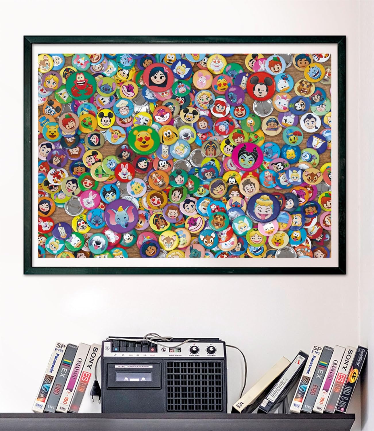 Puzzle Impossible Disney Emoji 1000 piezas Clementoni montado y enmarcado en negro sobre una pared, con la escena de chapas de emojis de personajes Disney; debajo, un radiocasete y cintas VHS en una estantería.