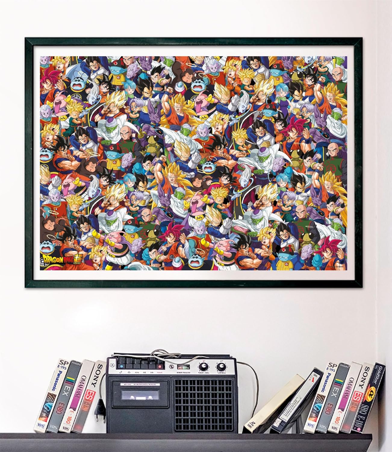 Puzzle Impossible Dragon Ball 1000 piezas Clementoni montado y enmarcado en negro sobre una pared, con el collage de personajes de Dragon Ball Super; debajo, un radiocasete y cintas VHS en una estantería.